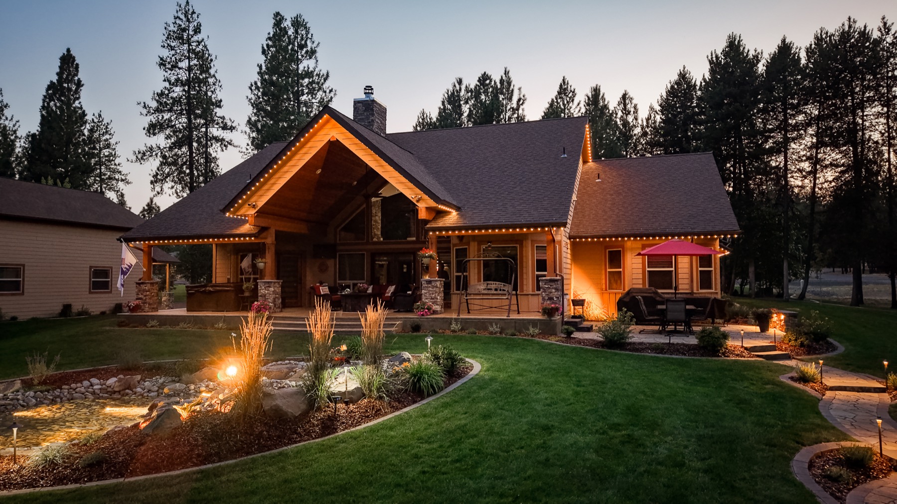 A backyard patio and landscaped pond lit with warm white trim and landscape lighting at dusk