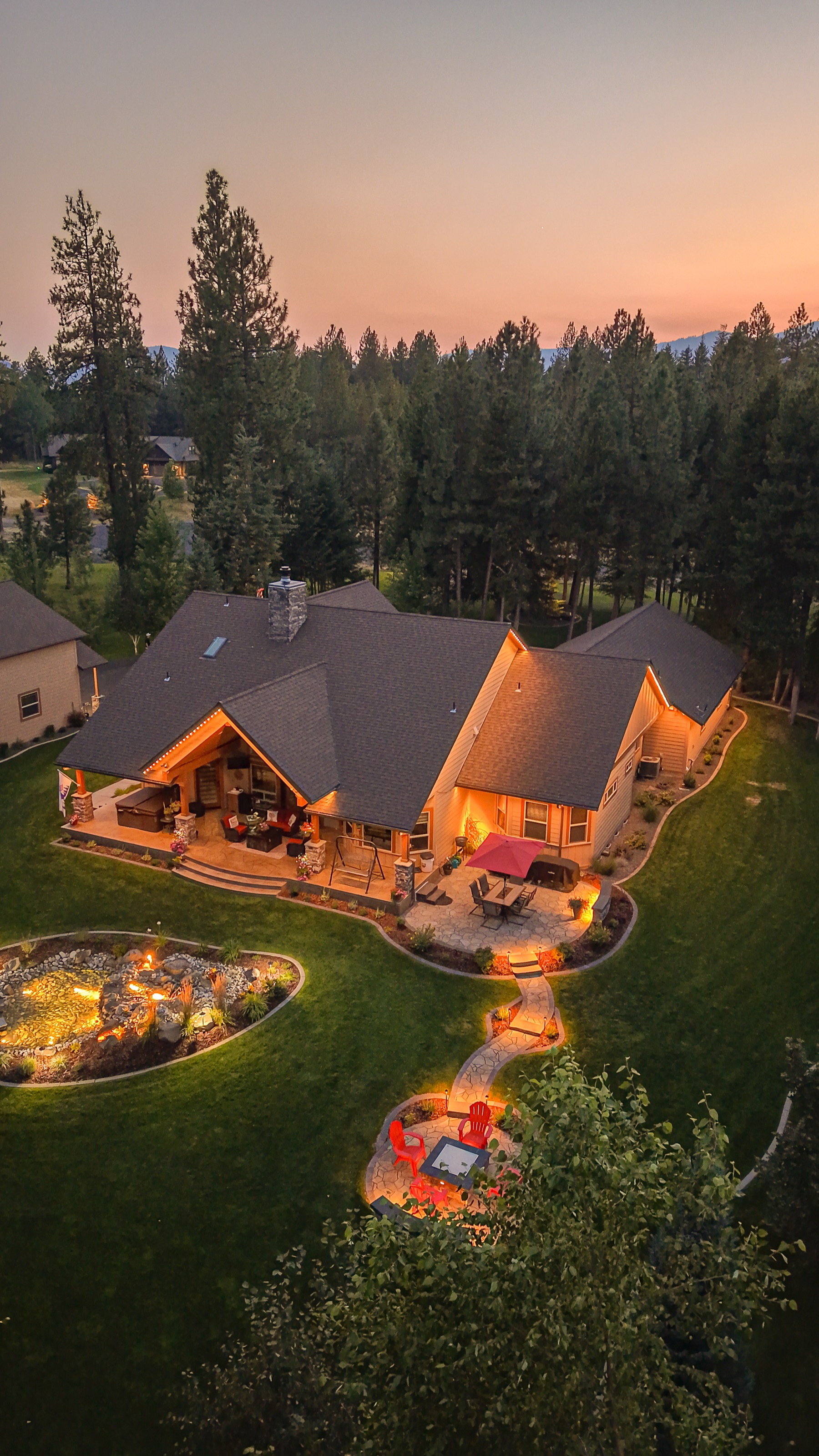 Aerial view of a home with a lit fire pit seating area and illuminated roofline at dusk